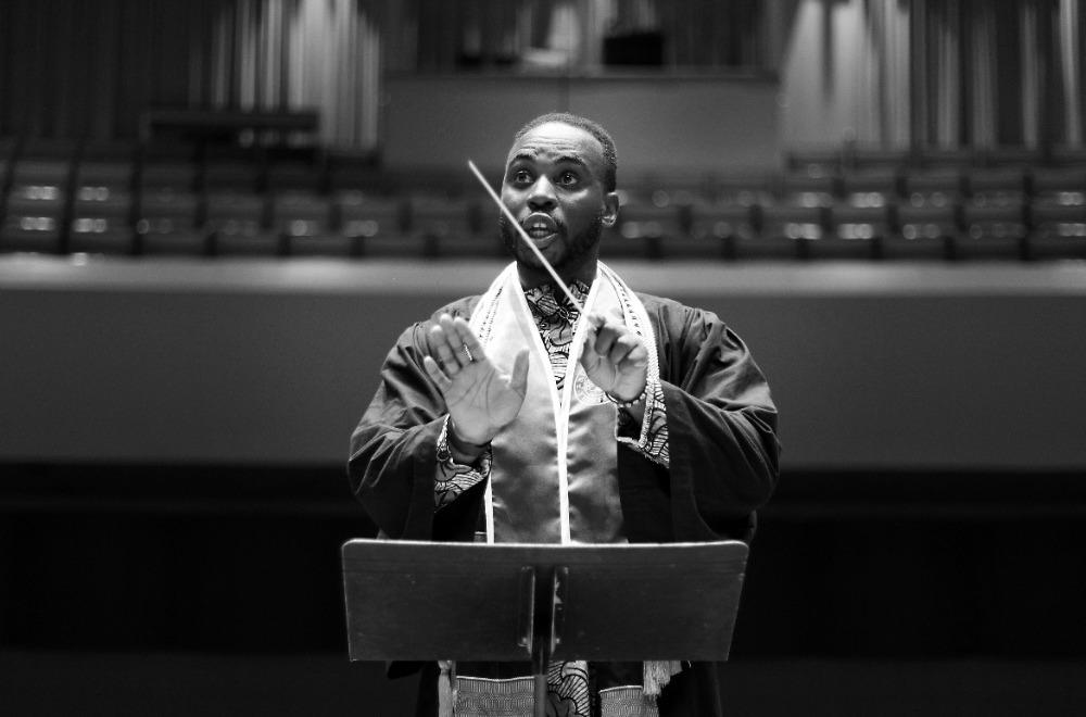 一张黑白照片，一位黑人男学生指挥戴着毕业礼服指挥合奏.